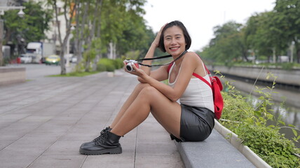 Fototapeta premium A cheerful Asian woman with short hair wearing a leather mini skirt and combat boots sits outdoors by a canal, smiling and holding her small digital camera
