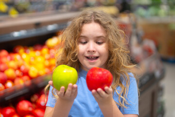 Fototapeta premium Kid holding a fresh apple in fruit store. Cute child holdi apple in fruit market. Red and green apples in kids hand. Apples in kids hand. Child face close-up. Child choosing a apple.