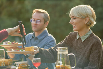 Caucasian middle aged woman smiling while receiving plate of food from another during outdoor meal, man sitting beside her holding plate and watching interaction