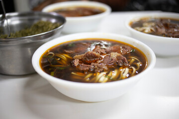 A steaming bowl of beef noodle soup featuring tender beef chunks, chewy noodles, and a savory, dark broth. Fresh herbs add color, while a side dish hints at a complete Asian-inspired meal.