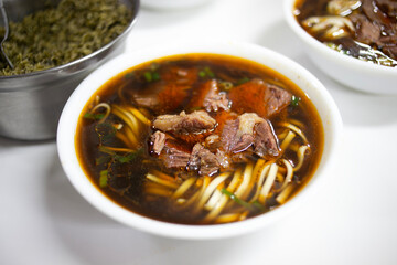 A steaming bowl of beef noodle soup featuring tender beef chunks, chewy noodles, and a savory, dark broth. Fresh herbs add color, while a side dish hints at a complete Asian-inspired meal.