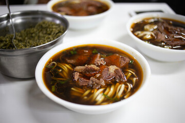 A steaming bowl of beef noodle soup featuring tender beef chunks, chewy noodles, and a savory, dark broth. Fresh herbs add color, while a side dish hints at a complete Asian-inspired meal.