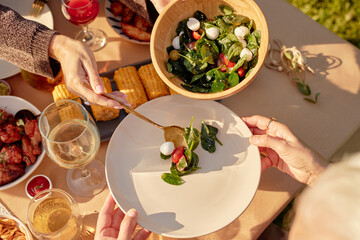 Senior Caucasian woman receiving fresh salad from middle aged Caucasian woman during outdoor meal, hands holding plate and serving spoon, visible corn and wine on table