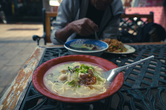 Close-up of a seafood soup noodles bowl on the table with a cropped image of a man eating fried rice in the blur background. 