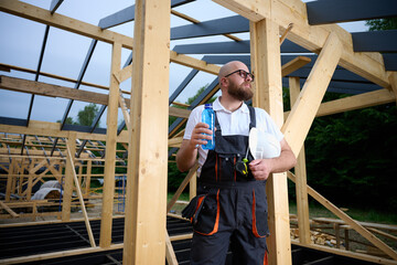 Construction worker taking a break and drinking water at wooden house frame construction site. Builder resting with hard hat and water bottle.