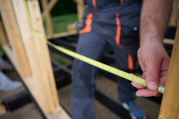 Construction worker measuring wooden beam with tape measure at building frame. Builder working on wooden house construction site outdoors. Crop view. Close up