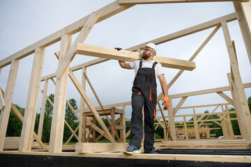 Construction worker carrying wooden beam on shoulder at building site. Carpenter in uniform and safety helmet working on timber house framework outdoors. Craftsmanship and construction work.
