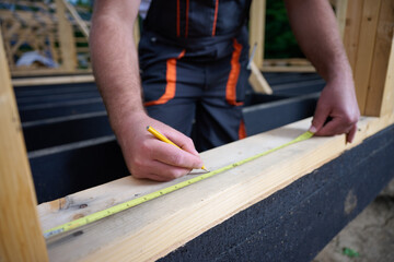 Close-up of construction worker marking wooden beam with pencil and tape measure at building site. Precision measurement during house frame construction.