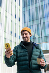 Smiling man using smartphone and holding coffee outside modern office building