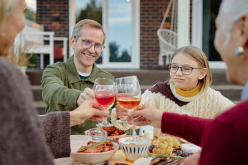 Caucasian middle aged man and Caucasian teenage girl sitting outdoors with family clinking glasses during meal, smiling and engaging in celebration at table with food