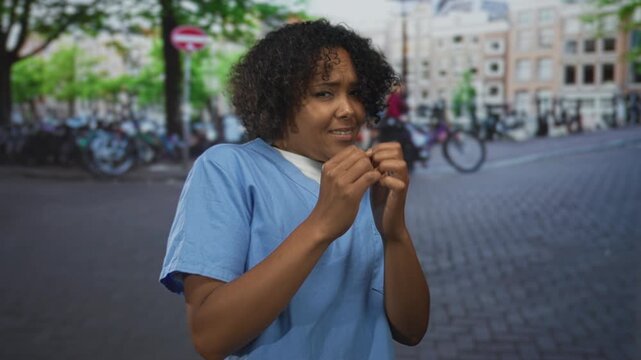 Woman covering face with hands on the city street while wearing blue scrubs and curly hair framing her head; anxiety.