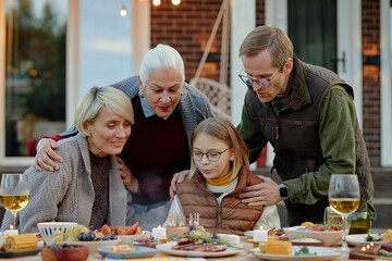 Caucasian senior woman, middle aged man, middle aged woman and preteen girl gathering around outdoor table celebrating birthday with cake and candles, sharing family moment