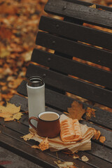 On the bench, there is a brown mug and a white thermos. A fresh bun is placed nearby, creating a cozy atmosphere.