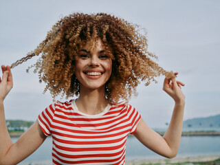 A joyful young woman with curly hair wearing a striped red shirt stands outdoors by a calm...