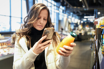 A young blonde white woman buys juice in a modern supermarket. She reads the label with her phone, scans it, and examines the ingredients. Healthy eating and healthy lifestyle.