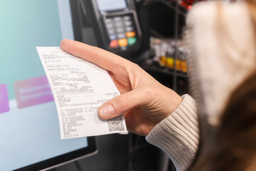 In a store, a woman closely examines her receipt while at a self-service checkout. The touchscreen display shows shopping options as she manages her budget.