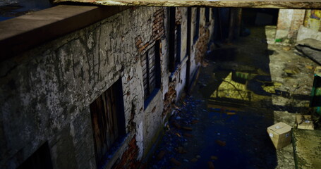 An old, deteriorating building features cracked walls and broken windows. Trash and old materials litter the ground, creating a sense of neglect and decay in low light.