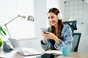 Young woman with headphones enjoying remote work, holding coffee and smartphone while smiling at...