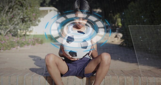Holding soccer ball, white-shirted boy sitting on brick ledge in backyard, showing digital overlay - Powered by Adobe