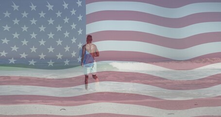 Fototapeta premium Wading man wearing trunks, carrying blue surfboard into ocean at beach, with translucent US flag