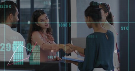 Two women in business suits shaking hands in lobby with clipboard laptop and digital charts