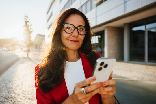 Confident woman in red jacket using smartphone outdoors in urban sunlight