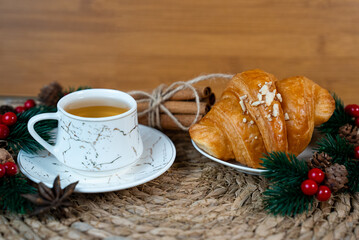 Cup of tea and croissant on woven mat with pine branches and Christmas decor. Warm cozy morning and holiday mood. 