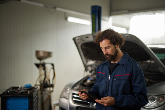 A car mechanic examines information on a smartphone while standing in a repair shop. A vehicle with an open hood is visible, indicating ongoing diagnostics. - Powered by Adobe