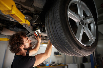 A car mechanic inspects and repairs a vehicle's undercarriage in a well-lit repair shop. Tools are scattered nearby as he focuses on his task.