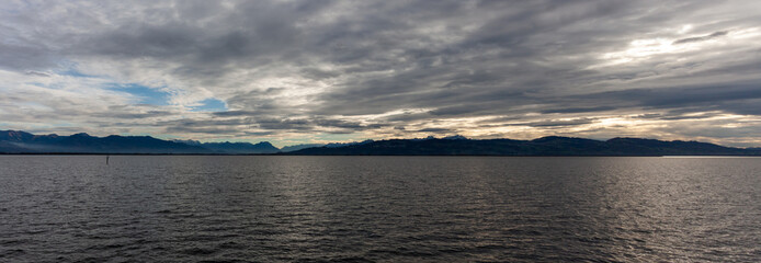 Dramatic panoramic view over the dark waters of Lake Constance (Bodensee) near Lindau, with the silhouette of the Swiss or Austrian Alps beneath a cloudy, moody sky