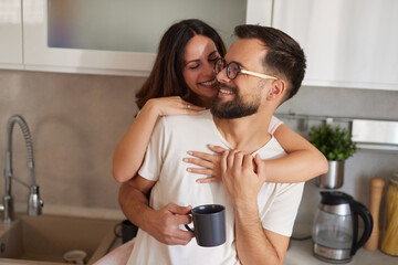 A couple starts their day in the kitchen, sharing laughter and smiles as one person holds a coffee cup while the other embraces them happily from behind, creating a warm atmosphere.