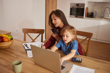 A mother balances work and family as she uses her laptop at the kitchen table. Her young child plays next to her, showing a cozy and productive family moment during the day.