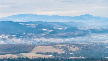 A tranquil winter panorama of Beskid Mały in southern Poland, where snowy meadows meet evergreen forests and rolling mountain hills under a serene, cloud-streaked sky.