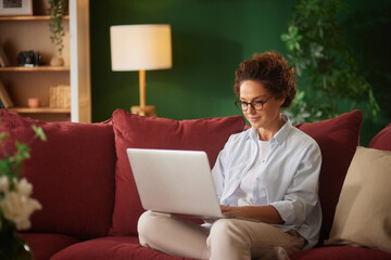 A woman sits on a red couch in a bright living room, focused on her laptop. She wears glasses and a...