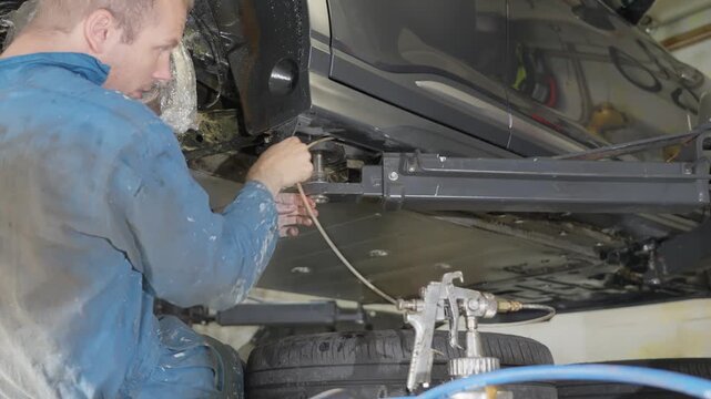 Man in coveralls applying lanolin rustproofing inside car sill and rocker panel cavities with long tube attached to pressurized spray gun