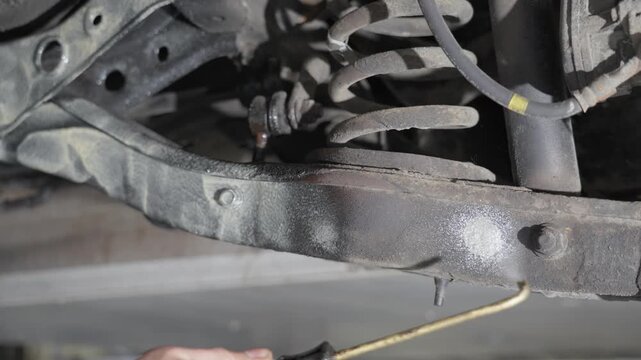 Person applying lanolin rustproofing under car, coating suspension and control arms with pressurized spray gun, close up