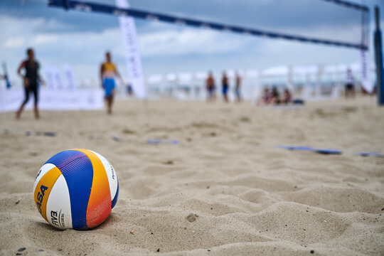 Beach volleyball tournament action with players competing on sandy court, net visible, professional FIVB ball in foreground