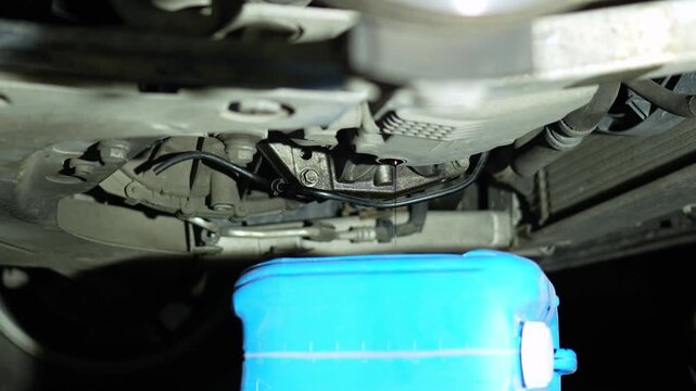 draining black used oil from a car engine into a blue canister. repair without a technical pit. home self-repair of the car. view from below the car. evening atmosphere, repair at night