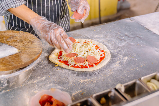 Chef assembling a pizza with fresh ingredients: dough, tomato sauce, mozzarella cheese, and sausage slices on a floured counter. Food preparation close-up