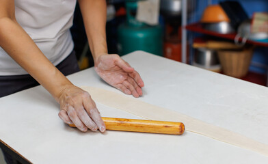 Close-up hand of asian young woman rolls bread dough with rolling pin on wooden cutting board for homemade bread cooking on table in kitchen, preparation ingredients process making food bakery baking.