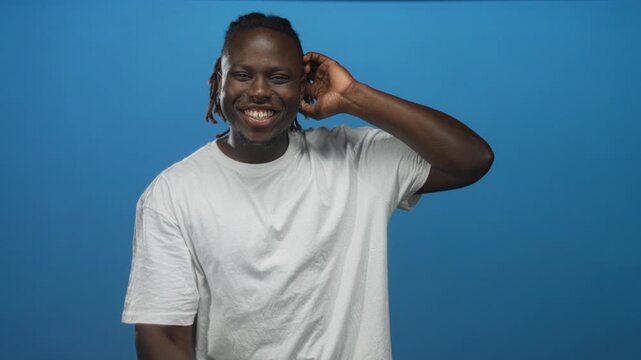 Man in white t shirt holding hand to ear and giving thumbs up in studio with solid blue backdrop; playful confidence.