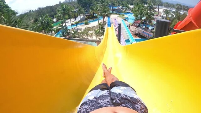 POV video: a rapid descent down a bright yellow water slide in a tropical water park. Summer vacation, speed and adrenaline; view of pools and palm trees. Great for travel, tourism, and lifestyle.