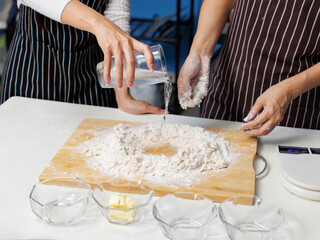 Close-up hand of asian woman baker wearing striped apron mixing pour flour different ingredients, kneading dough, process making homemade preparing bread pastry dough baking in bakery kitchen concept.