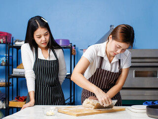 Two asian pretty young woman bakers wearing striped aprons knead dough on wooden cutting board mixing flour baking process different ingredients making homemade bread pastry in bakery kitchen concept.