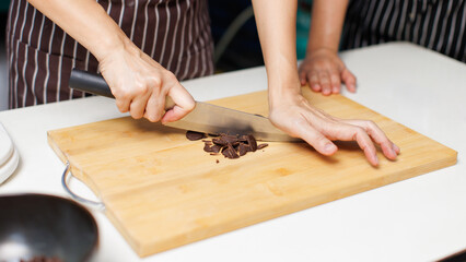 Close up hand of asian woman baker wearing striped apron chopping cutting chocolate bar with knife...