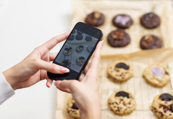 Hand of woman takes picture on smartphone freshly baked homemade dark chocolate chip soft cookies topping biscuits wafer marshmallow on baking paper sheet dessert snack bakery yummy food photo concept