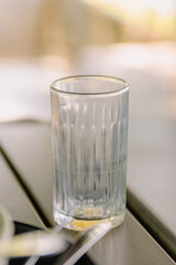 Simple, clear empty drinking glass with a subtle ribbed texture, placed on a dark table with a soft, blurred outdoor background