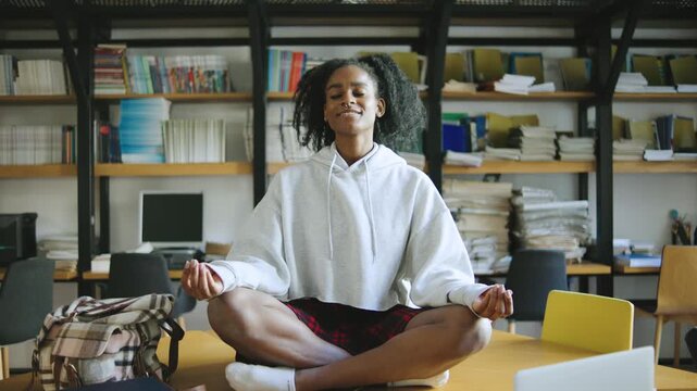 Young African American woman practices mindfulness and meditation in a quiet study area surrounded by books and computers during a calm afternoon