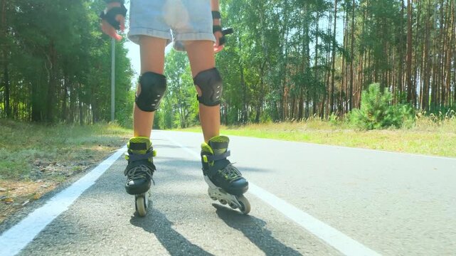 Child girl in casual clothes roller skating on road in forest on sunny summer day.