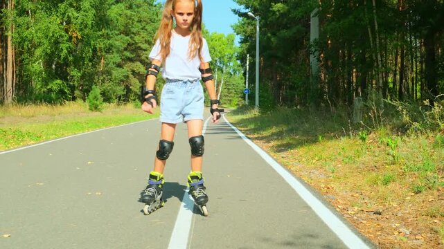 Child girl in casual clothes roller skating on road in forest on sunny summer day.